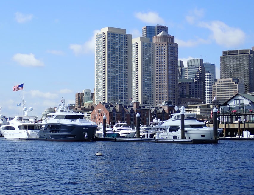 Luxury boats docked in a harbor with a city skyline rising behind them under a blue, partly cloudy sky.