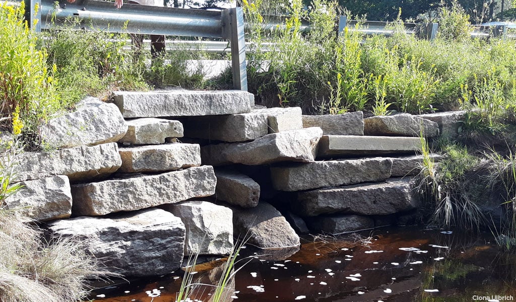 Riverbank view of culvert made from rectangular stone slabs with roadway guardrail above it.