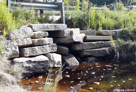 Riverbank view of culvert made from rectangular stone slabs with roadway guardrail above it.