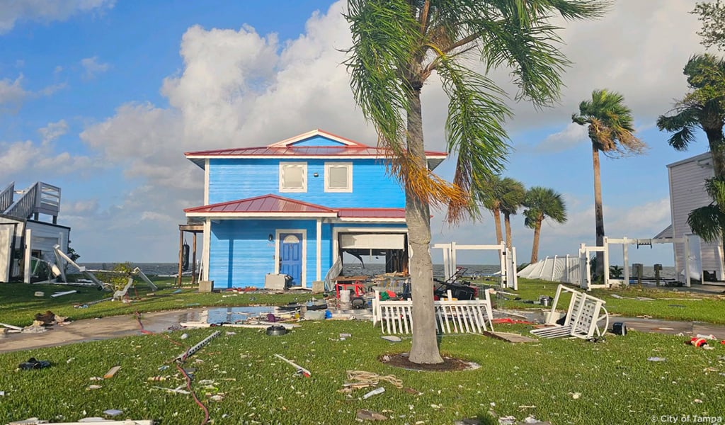 A bright blue beach house with storm damage and debris along a coastline.