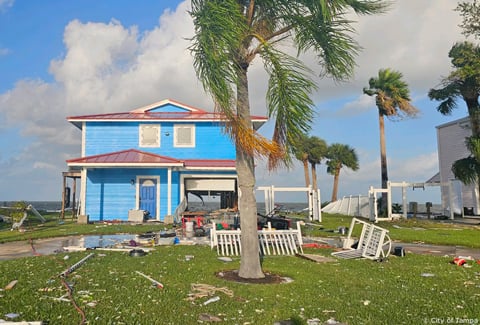 A bright blue beach house with storm damage and debris along a coastline.