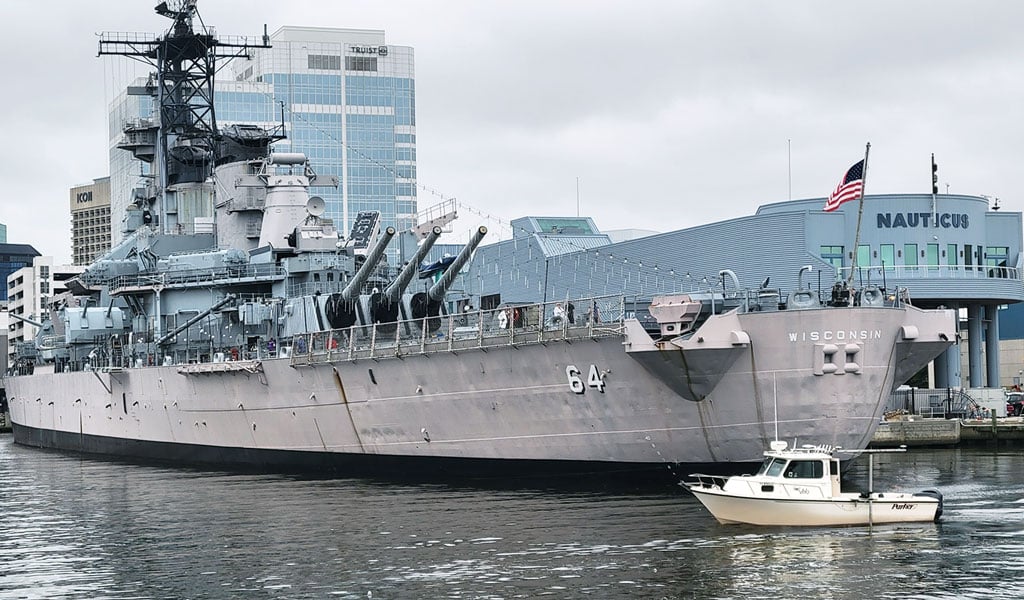 A VHB survey boat passes a large gray battleship, the USS Wisconsin, in a harbor near a waterfront city.