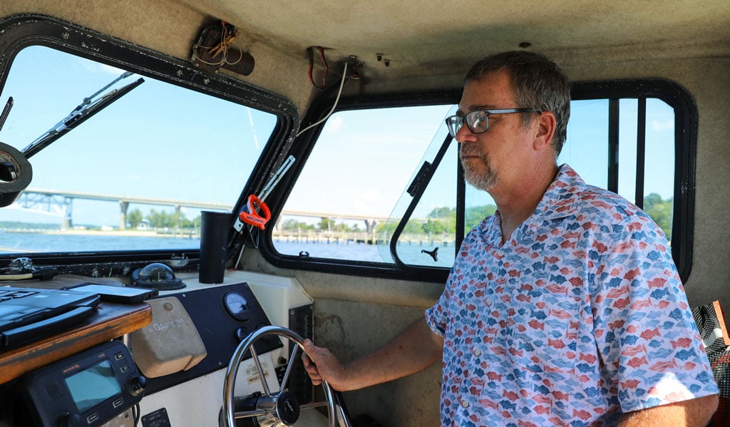 A man drives a boat on the water with a bridge visible through the windows.