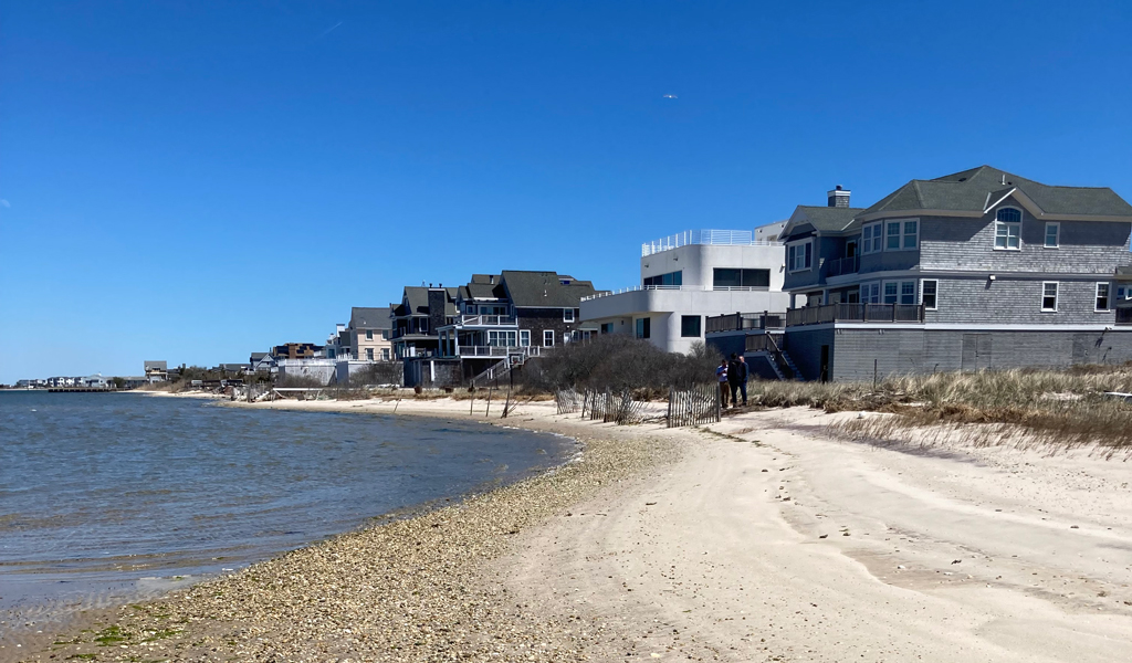 A coastline featuring a narrow strip of beach, with a row of homes sitting about away from the shoreline. 