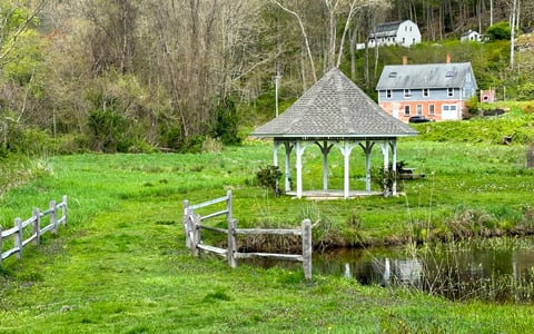 A serene rural landscape featuring a gazebo by a pond, with lush green grass and a wooden fence in the foreground.