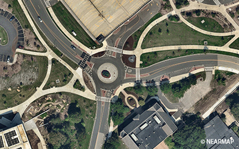 Aerial view of a roundabout with pedestrian crossings and surrounding roads, featuring vehicles and signage, set in a landscaped urban area.