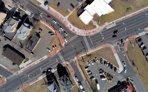 Aerial view of an intersection with multiple crosswalks and traffic lanes, vehicles parked along the curbside, and patches of green areas beside the roads.