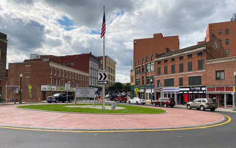 A view of a small urban center with a roundabout featuring an American flag and a historical marker. 