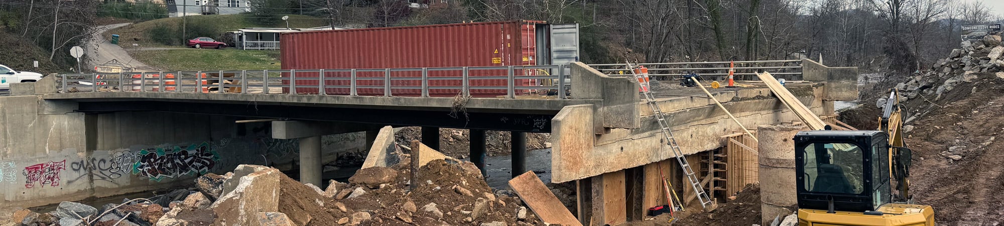 Hurricane-damaged bridge in North Carolina with collapsed roadway, debris, and excavator working on recovery efforts.