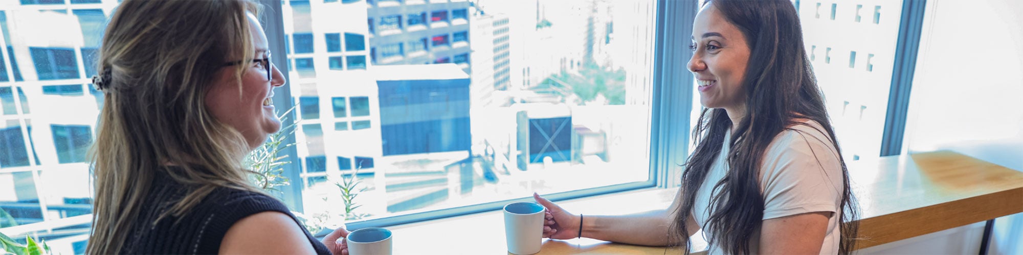 Two employees seated near an office window chatting informally over a cup of coffee.