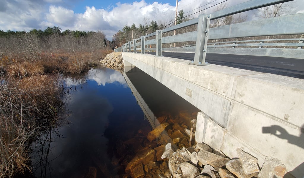 Concrete bridge over a clear stream with visible rocks underneath, surrounded by dried vegetation under a partly cloudy sky.