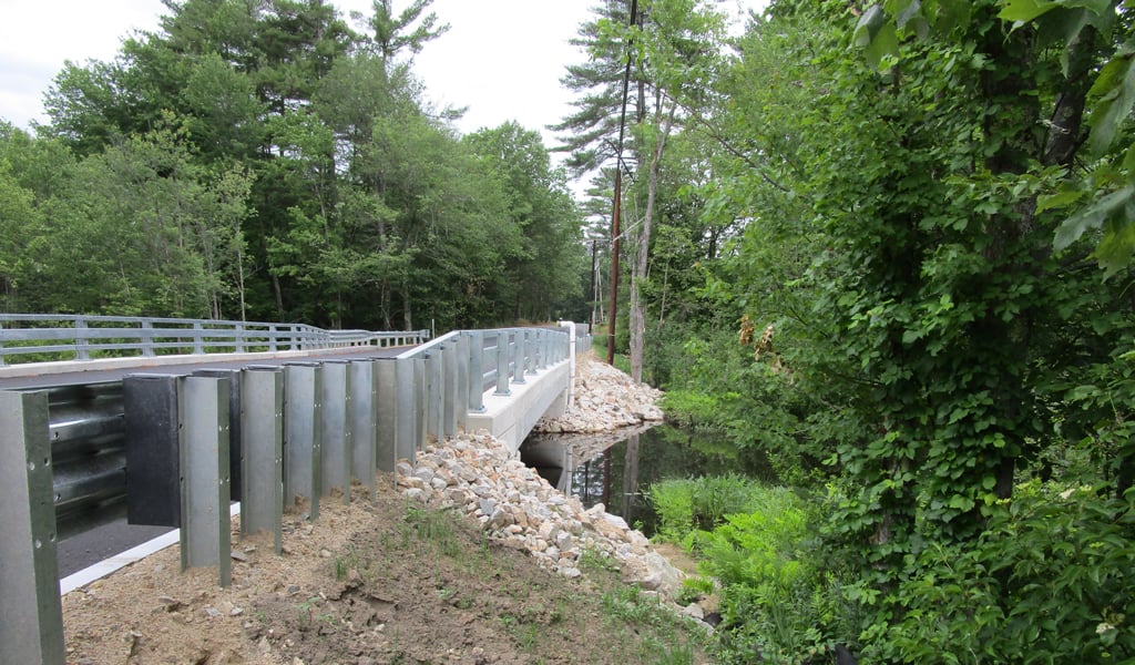 View of a newly constructed bridge surrounded by lush greenery, with a clear sky above and a stream flowing underneath.