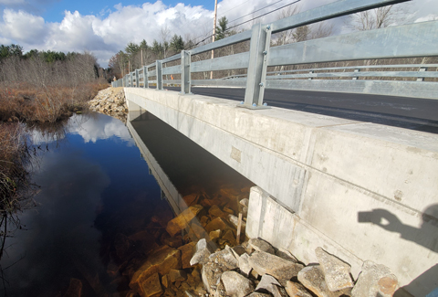 Concrete bridge over a clear stream with visible rocks underneath, surrounded by dried vegetation under a partly cloudy sky.