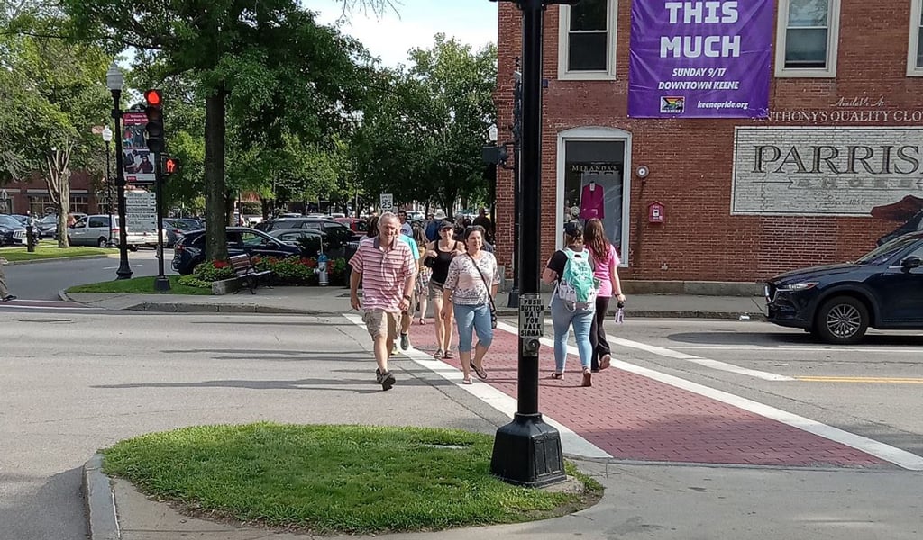 Pedestrians crossing a street in downtown Keene, NH near a signpost and Parrish Shoes wall mural.