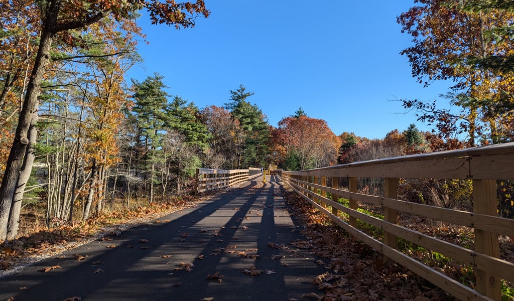 Autumn scenery along a paved path with a wooden fence on one side, surrounded by trees with fall foliage under a blue sky.