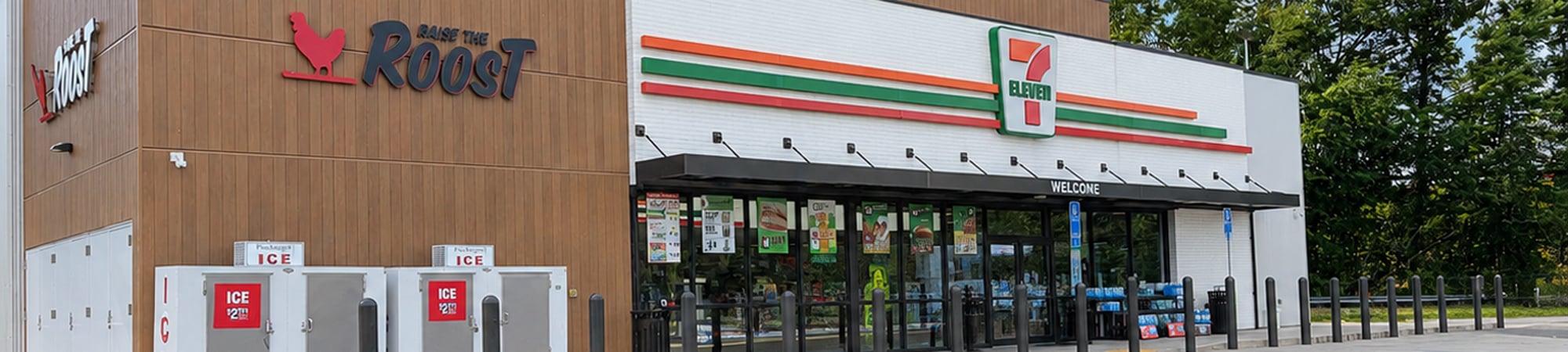 The front entry of a 7-Eleven Raise the Roast store with grey bollards in the foreground. 