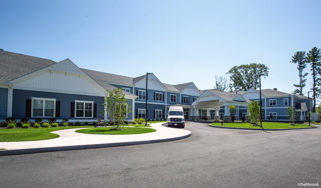 Two-story senior living residence with a van at the covered front entrance on a sunny day.