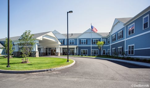 Blue two-story senior living residence with a covered entrance, circular driveway, lawn and flagpole on a clear, sunny day.