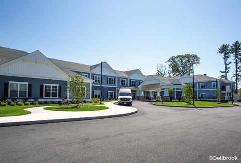 Two-story senior living residence with a van at the covered front entrance on a sunny day.