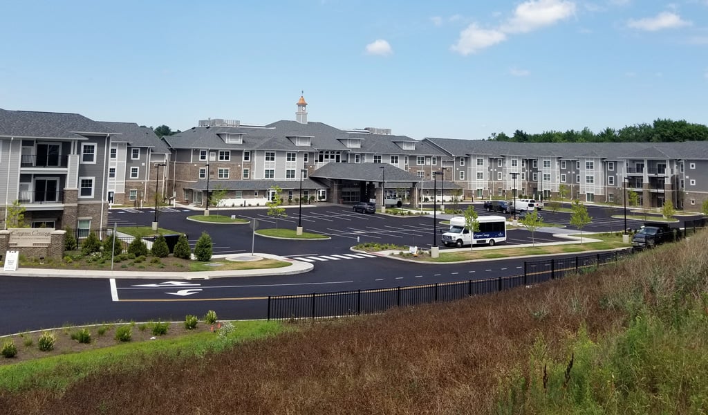 Modern gray three-story senior living facility with a large parking lot under a bright blue sky with scattered clouds.