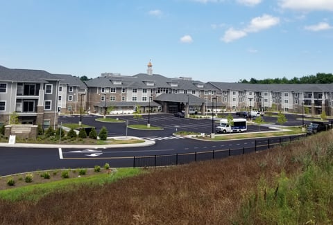 Modern gray three-story senior living facility with a large parking lot under a bright blue sky with scattered clouds.