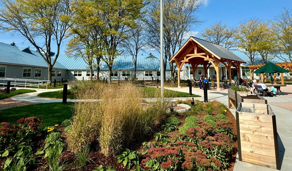 Senior living courtyard with pavilion, flagpole, accessible walkways, and landscaped gardens with seating on a sunny day.