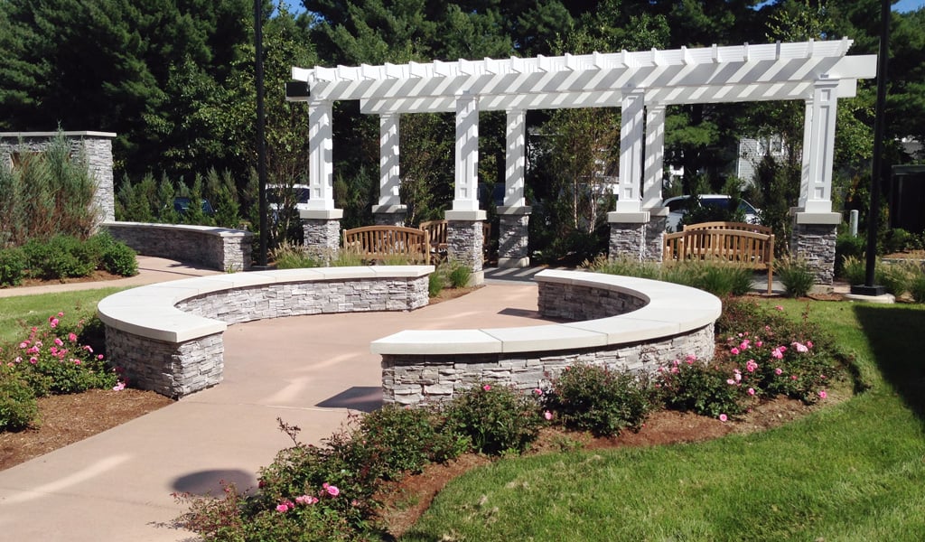 Tranquil garden courtyard with green grass surrounding a circular area with stone walls and an arbor in the background.