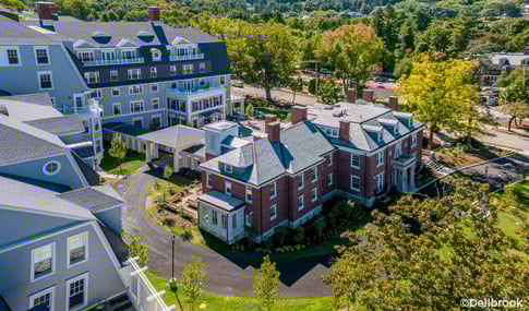 Aerial view of a multi-story senior living complex with brick and gray buildings surrounded by trees with Boston in the distance.  
