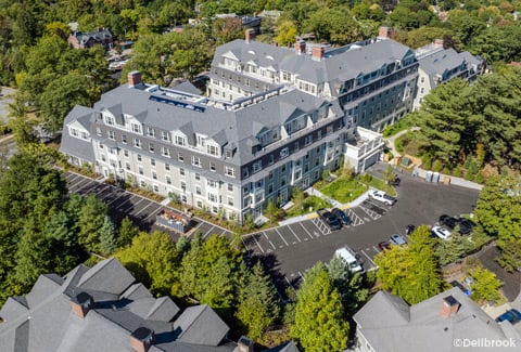 Aerial view of a multi‑story senior living complex with parking lots, trees, and a town and lake in the background.