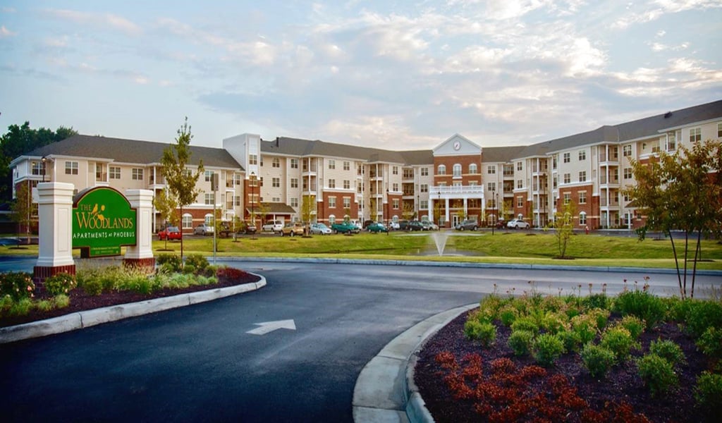 A 4-story senior living facility with a green lawn, fountain and landscaped roadways in the foreground. 
