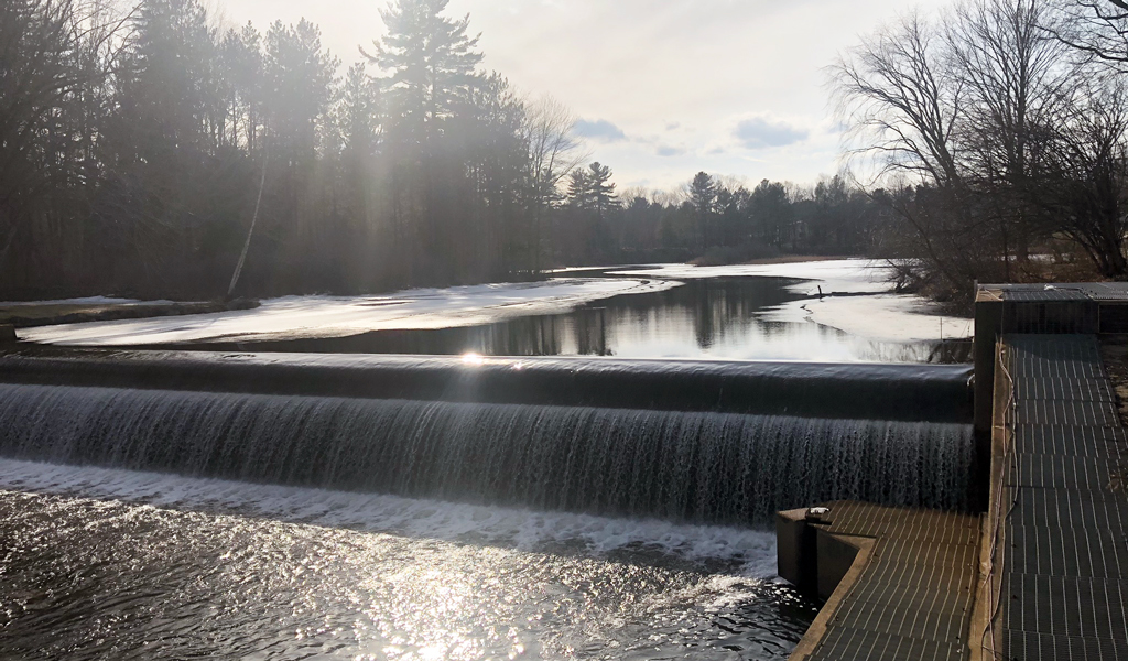 A river with a smooth waterfall flowing over a dam, surrounded by trees under a partly sunny sky. 