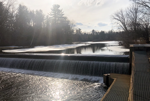 A river with a smooth waterfall flowing over a dam, surrounded by trees under a partly sunny sky. 