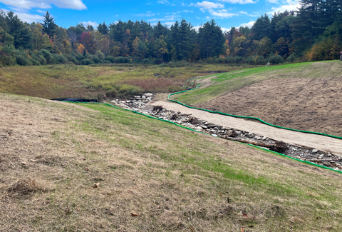 Landscape with a narrow, rocky water stream, surrounded by grass, trees, and a partly cloudy blue sky.