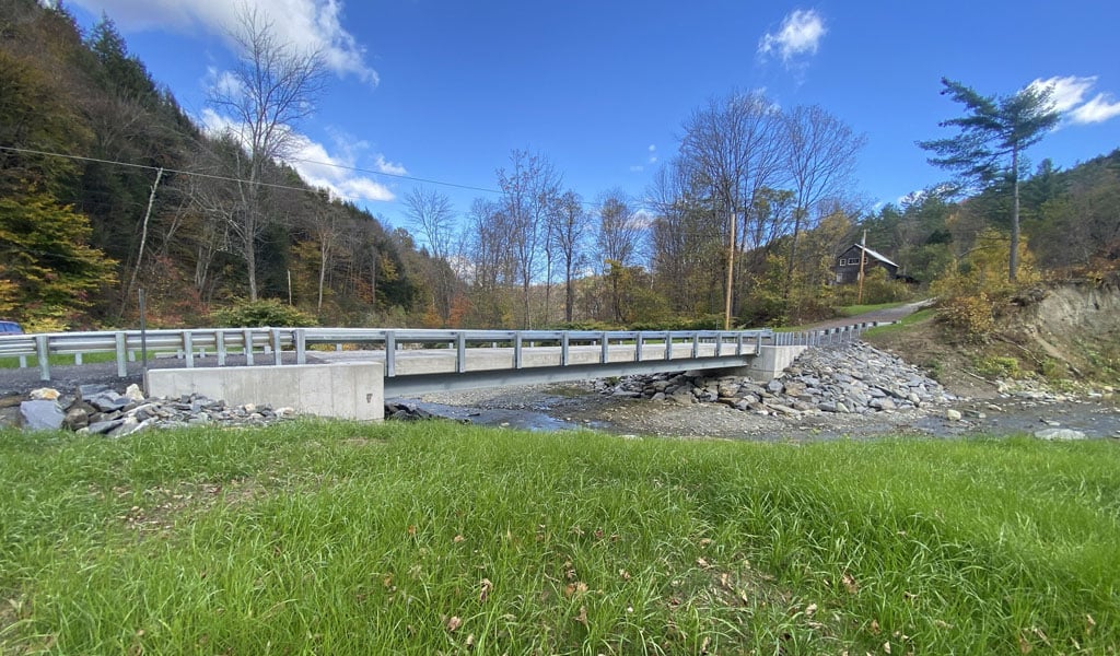 A sturdy bridge crosses a small, rocky stream, surrounded by green grass and bare trees. 