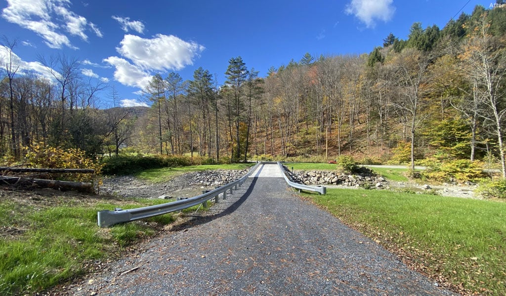 A narrow gravel road with metal railings leads through a scenic landscape of trees with autumn foliage. 