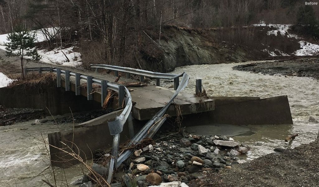 A small, damaged bridge with twisted metal railings spans a rocky stream surrounded by bare trees and patches of snow. 