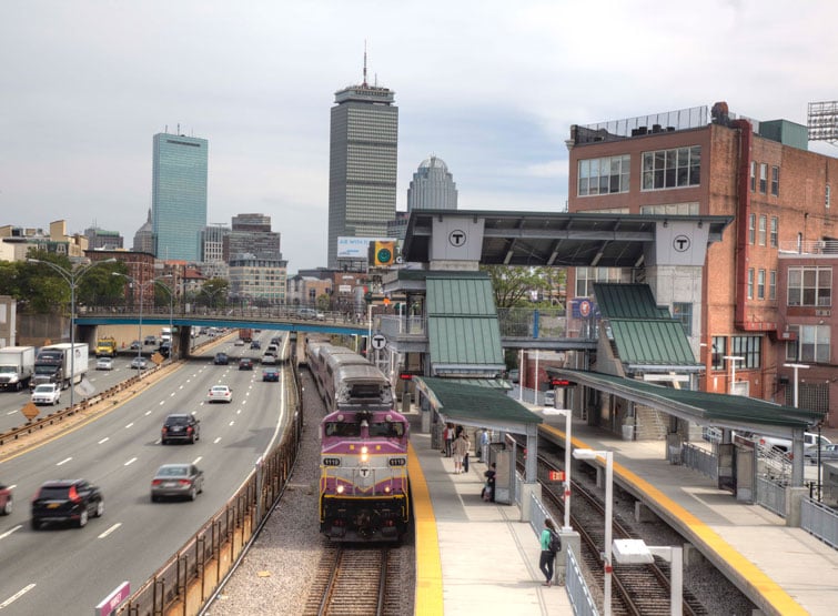 A train at a rail station next to a road with cars. 