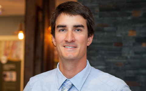 Person smiling in an office environment with a casual blue shirt and tie, standing in front of a rustic wooden beam and a dark stone column.
