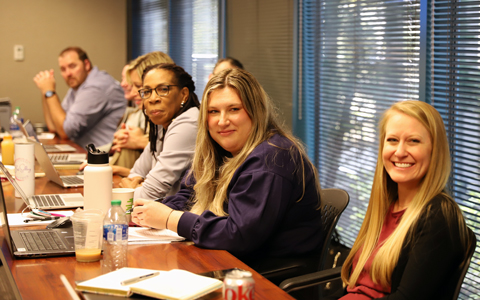 Alex DeYoung smiles in an office setting at a conference table with colleagues