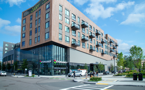 Modern urban building housing a Stop & Shop store on the ground floor, with a mix of glass and wooden facade, set against a backdrop of a clear blue sky with scattered clouds.