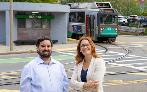 Two individuals smiling in front of a Heath Street station with a moving tram labeled 'E Line'.