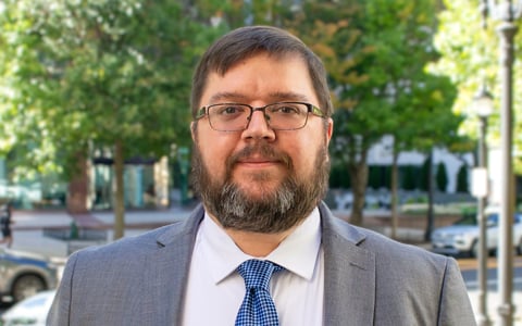 Man wearing glasses smiling in a grey suit with a blue checkered tie surrounded by Atlanta’s green trees.