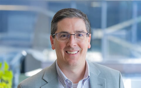 Chris Fronheiser wears glasses and a grey blazer in an office setting. 
