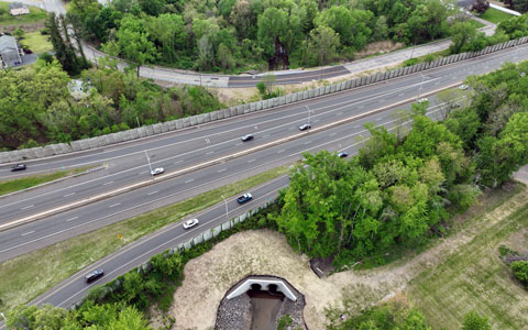 Aerial view of a highway with four lanes showing moderate traffic, surrounded by lush greenery and a small tunnel passing under one side of the road.