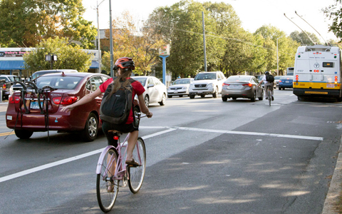 A woman wearing a bicycle helmet and riding a bike in a bike lane uses hand signals on a busy road.