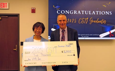 Two individuals holding a large ceremonial check from VHB, Inc., celebrating CSTI Graduates under a "CONGRATULATIONS" banner