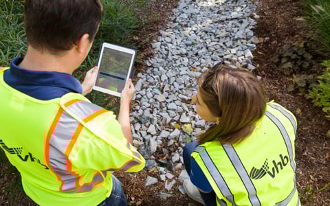 Two individuals in high-visibility vests are crouched over a tablet by a gravel path, examining a digital map.