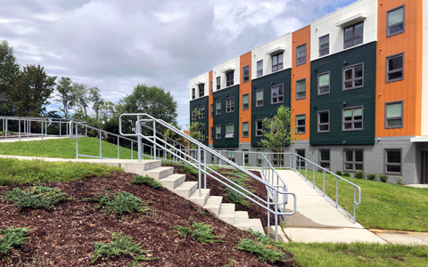 A white, green, and orange student housing building is bordered by accessible paths and green space. 