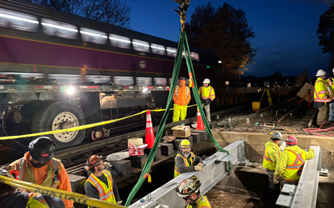 Workers in high-visibility clothing install large equipment near a commuter train during nighttime hours.