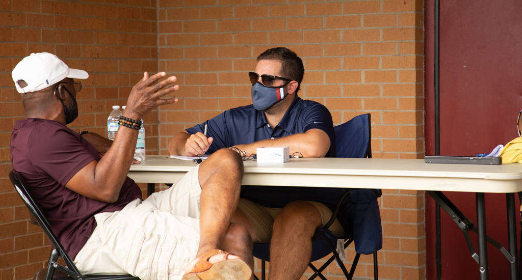 Two men wearing masks sit at a folding table outside a brick building while one man takes notes